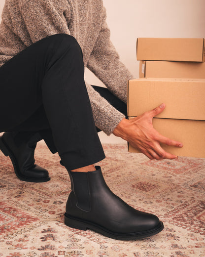 Person wearing black boots and holding cardboard boxes on a patterned rug.