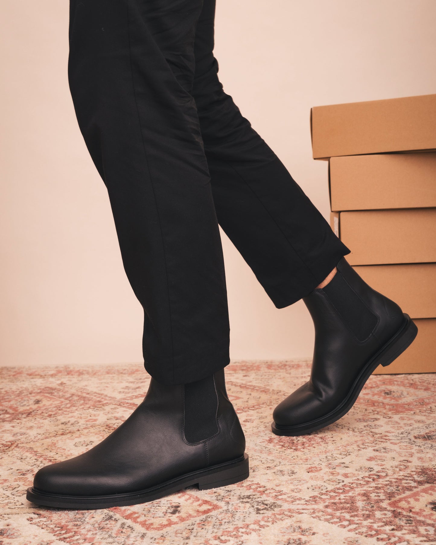 Person wearing black Chelsea boots with a beige wall and stacked boxes in the background